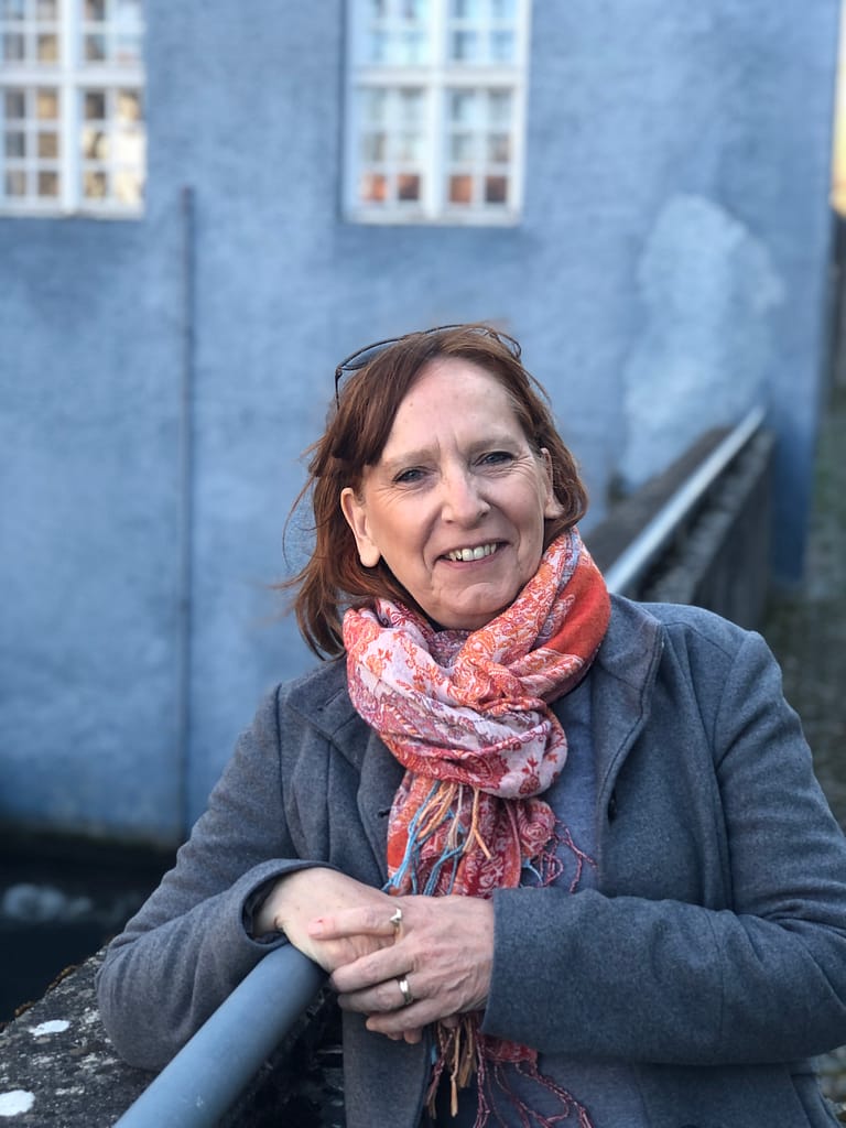 Eine rothaarige Frau in einem grauen Mantel und einem gemusterten Schal steht im Freien und lehnt sich an ein Geländer vor einem hellblauen Gebäude mit weiß gerahmten Fenstern.